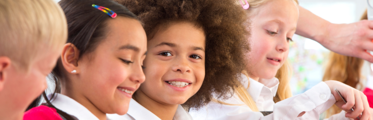 Four children in school uniform working at a table, one looking towards the camera smiling